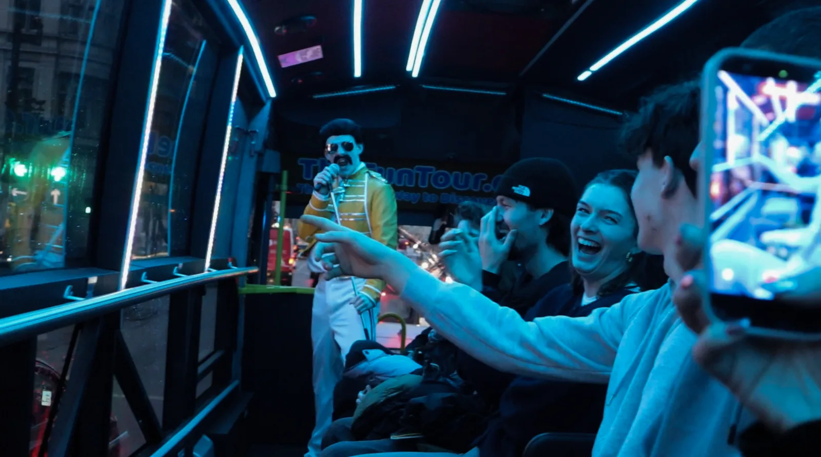 A rock legend in a yellow jacket performing at the front of the bus while guests laugh and film